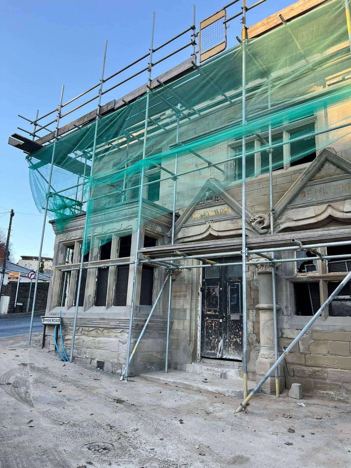 Victorian stone building with carved pediment gables and a panelled front door, wrapped in scaffolding and green safety netting during restoration