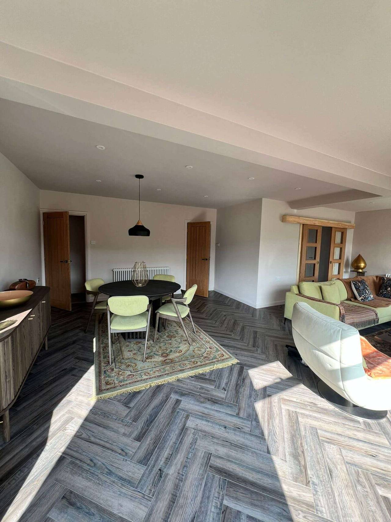 Bright open-plan living and dining room with grey herringbone flooring, oak doors, a round dining table with sage-green chairs and a sofa