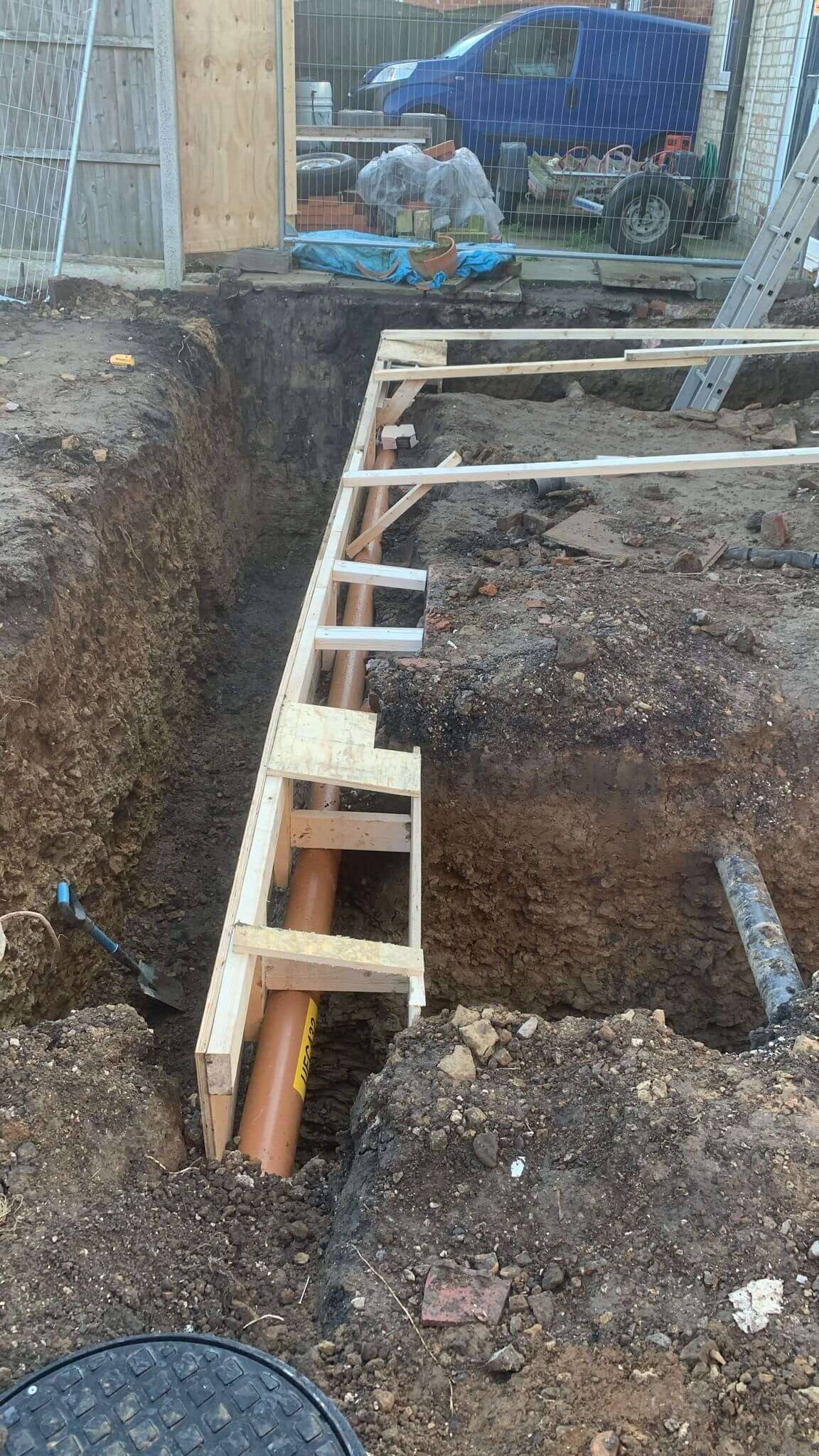 Extension foundation trench with timber shuttering and a new orange drainage pipe running alongside